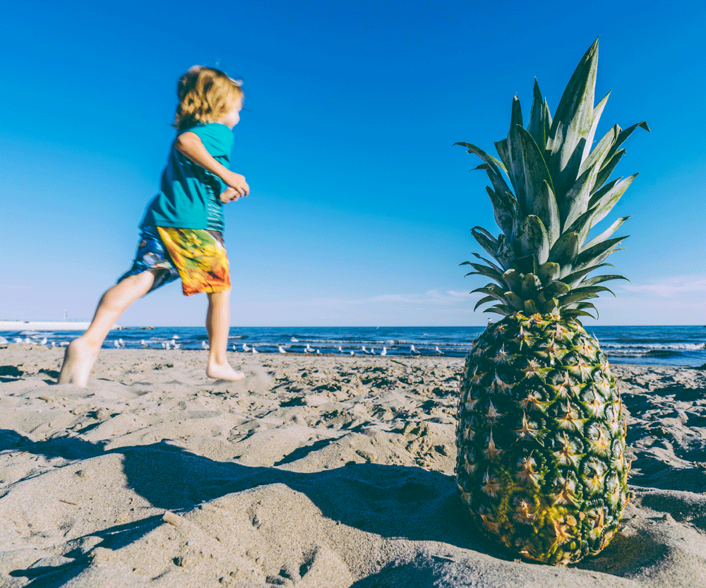 A young boy runs past a pineapple on the beach, packing your own snacks is one of our best tips for taking a family vacation on a budget.