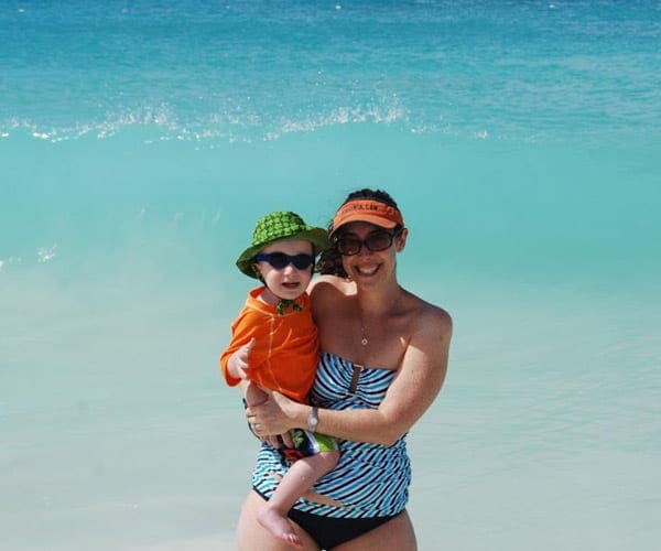 A mom holds her young child, both are smiling, in swim ware on the beach in Aruba.