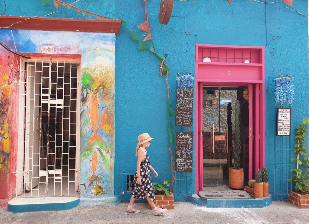 Girl in dress and straw hat in front of blue and pink shops in Cartagena, Colombia.