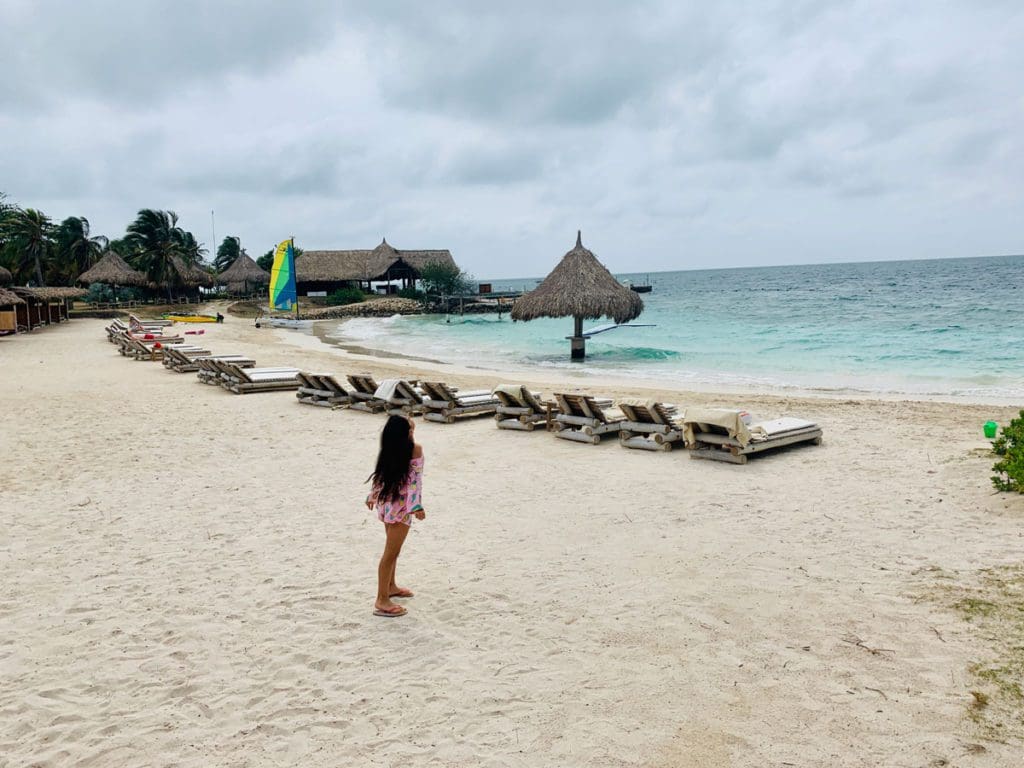 A young girl looks up a the sky while exploring a beach at Punta Faro, rows of beach loungers and the ocean are seen in the distance.
