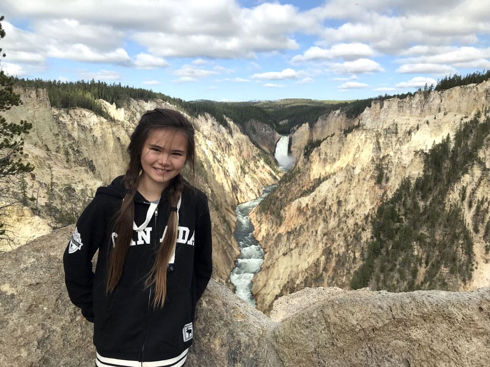 Girl in front of a waterfall while hiking in Yellowstone National Park, one of the best Summer Vacation Ideas in the U.S. for Families.