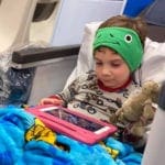Child on airplane seat with blue blanket, stuffed animal and iPad in pink case with green headphones