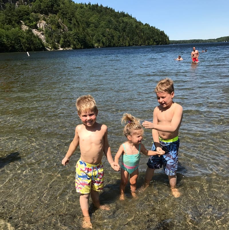 Three young kids stand holding hands in the water. Going to the beach is one of the best things to do while exploring Acadia National Park with kids.