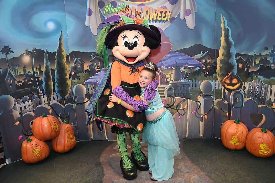 A young girl wearing a Princess Jasmine costume hugs Minnie Mouse decked out in her Halloween ensamble.
