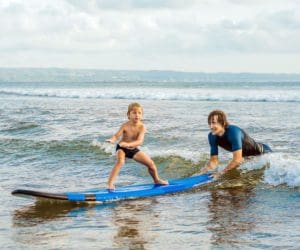 A young boy learns to surf in Hawaii, with an instructor behind him helping balance the surf board.