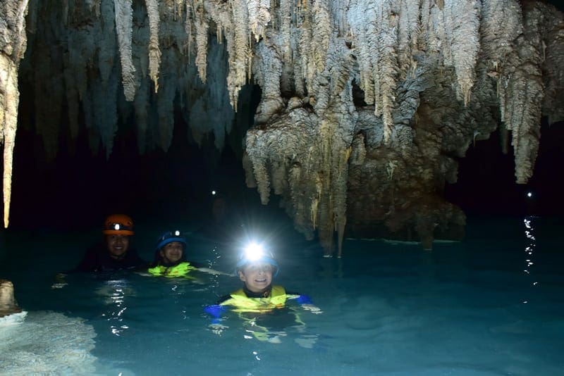 Family of three with flashlights swimming in Rio Secreto underground cave, one of the best things to do in Playa del Carmen with kids.