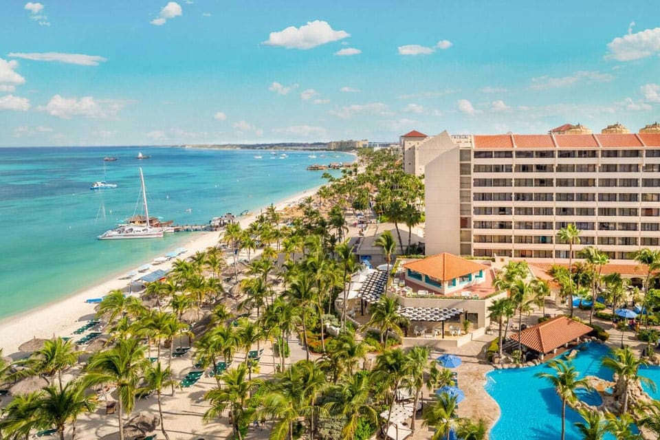 Aerial view of Barceló Aruba, featuring a blue pool, green palms, and large buildings. Affordable accommodations is is one way to have an affordable Aruba family vacation.