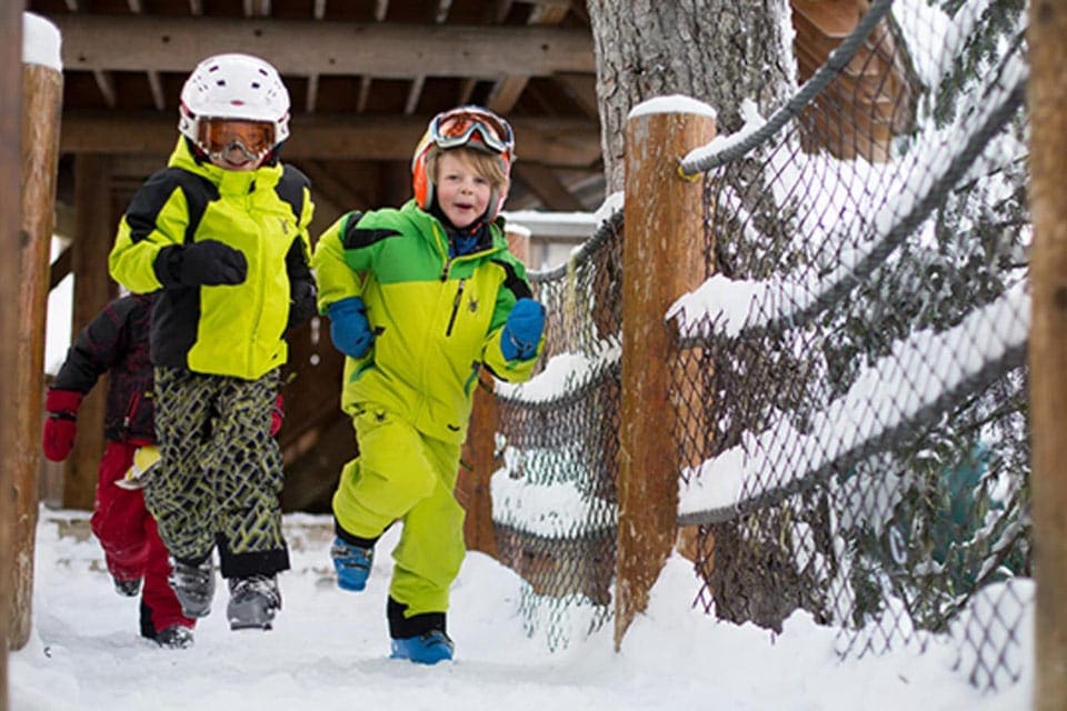 Two kids wearing bright winter gear run along a trail at Magic Castle and Tree Fort.