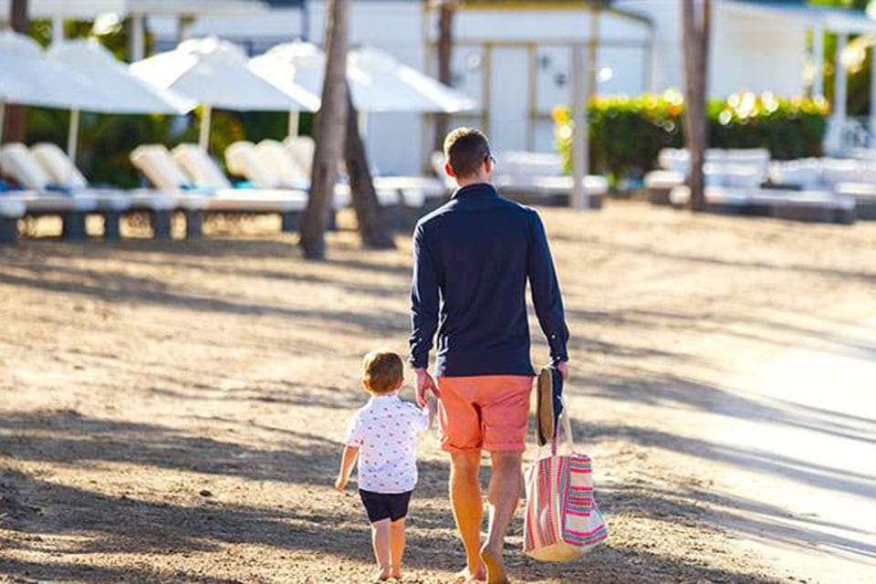 A dad holds the hand of us son, which a beach bag in the other hand, while meandering the sandy beach.