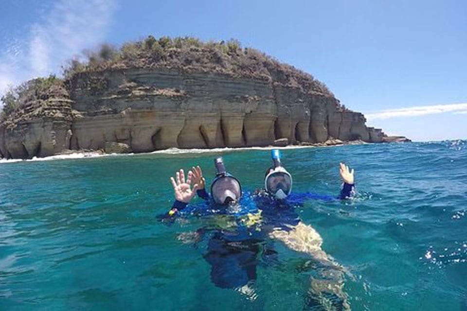 Two people wave while snorkeling off-shore from the Pillar of Hercules.