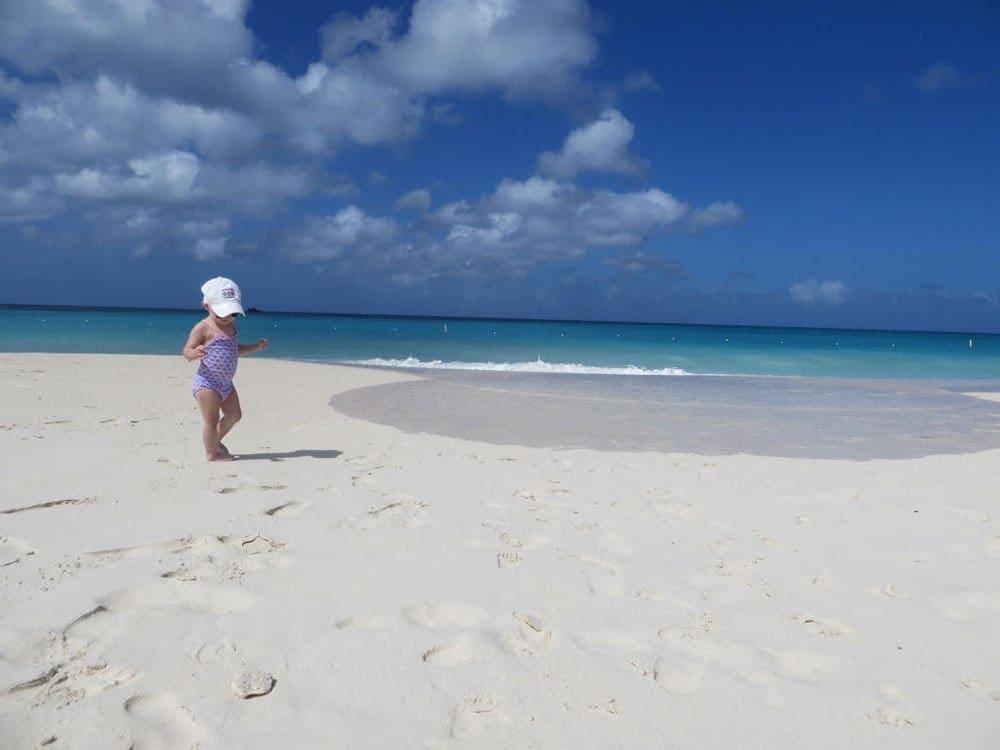 A young baby walks along a white sand beach in Aruba with blue skies behind her.
