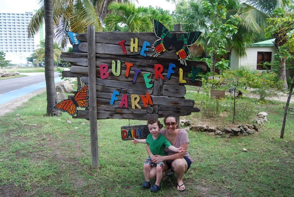 A mother and child kneel by the sign for the Butterfly Farm in Aruba.