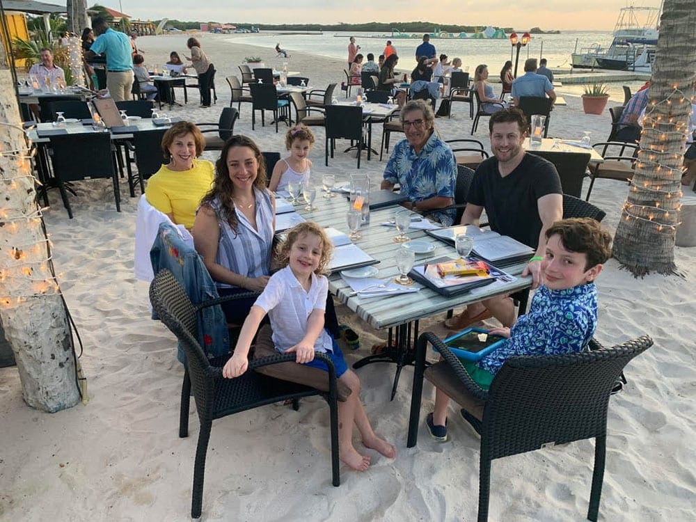 A family enjoys a meal on the beach in Aruba at sunset. Planning transportation ahead of time for meals is one way to have an affordable Aruba family vacation.