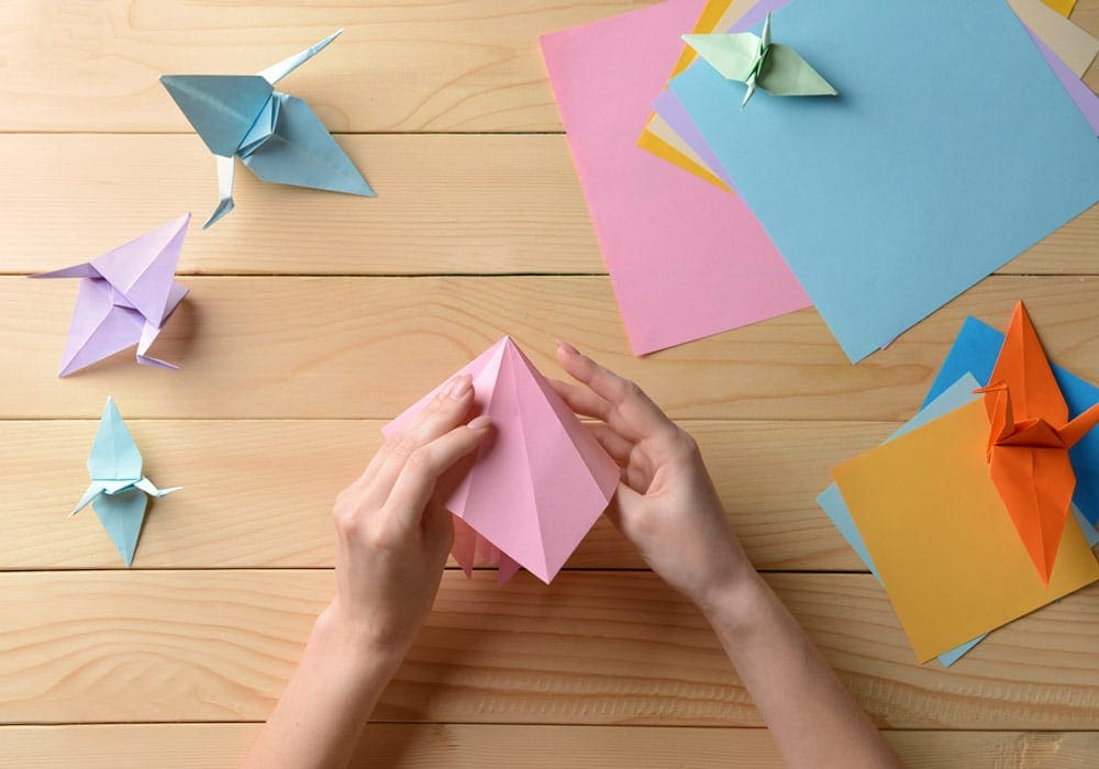 A pair of white hands folds pink paper while constructing an origami crane. Getting creative is a fun way to take a virtual vacation to China.