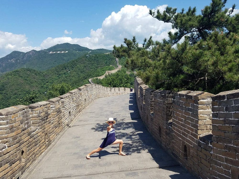 A young girl stands on the Great Wall of China with legs stretched and gives the camera a thumbs up.