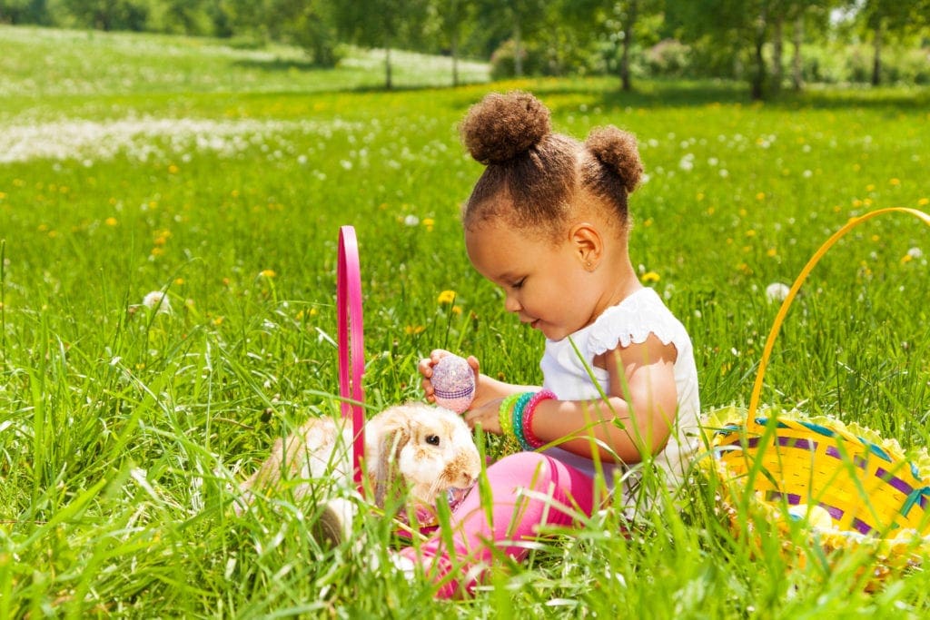 A young black girl sits in a grassy area with a yellow Easter basket, fluffy bunny, and holding a purple Easter egg.