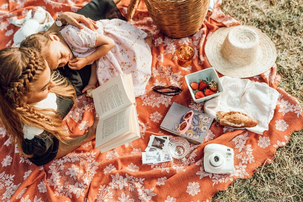 Mother and daughter snuggle reading a book on a picnic blanket. Reading together can be a wonderful way to connect on your virtual vacation to Norway.