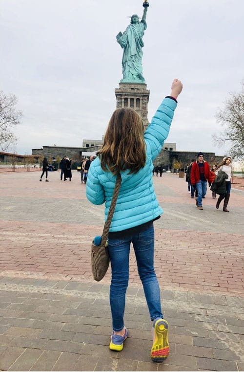 A young girl stands with her fist in the air facing the Stature of Liberty in the background.