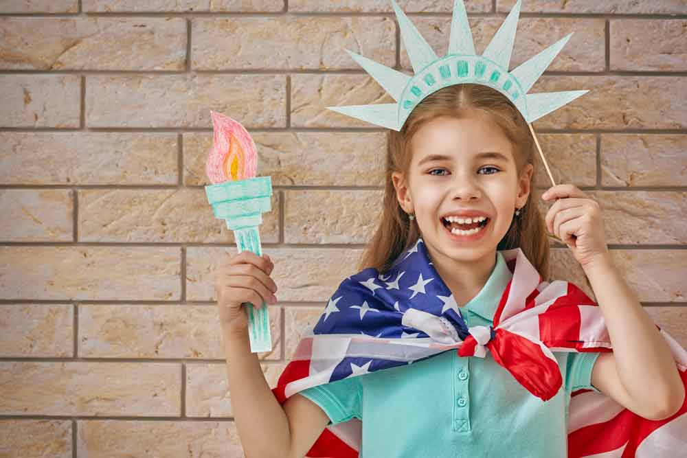 A girl poses with Statue of Liberty craft hat on her head and torch in her hand, while wearing the American flag as a cape.