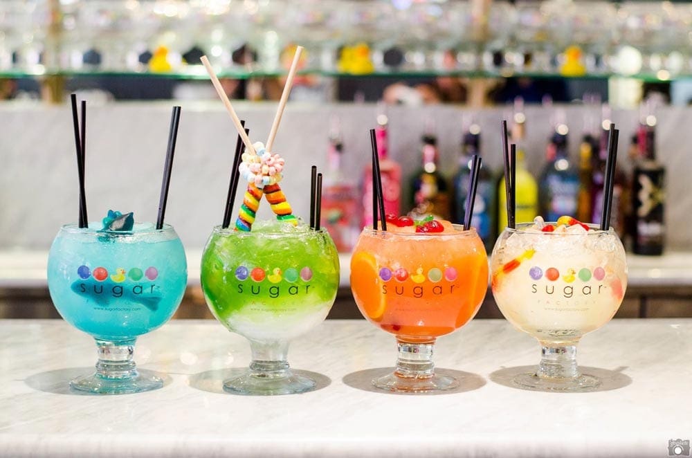 Four drinks, one in blue, green, orange, and white, respectively, line a bar at Sugar Factory.