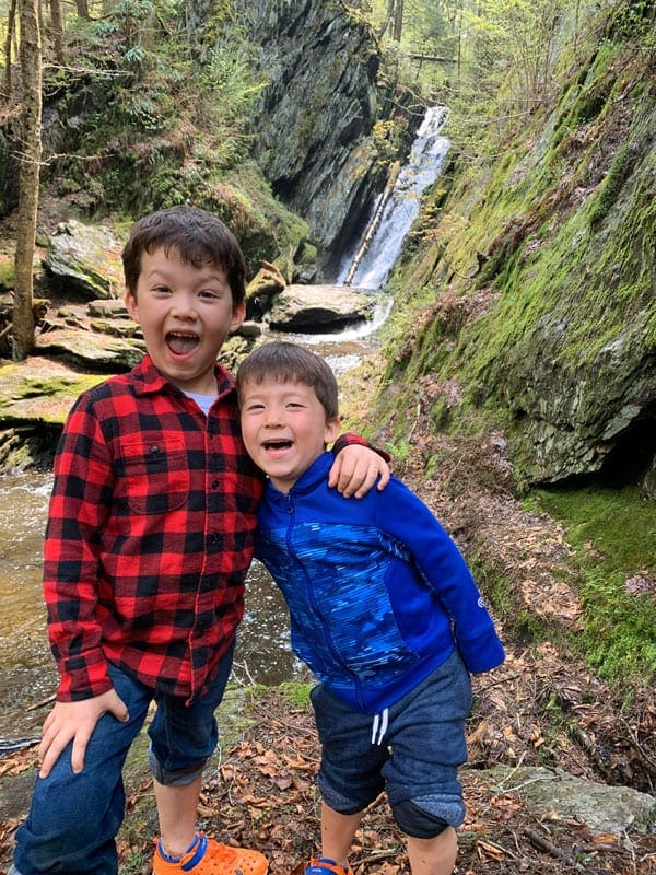 Two brothers stand together laughing in front of a waterfall in the forest, while exploring the Great Barrington.