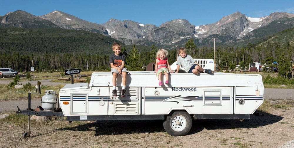Three kids sit on top of a pop-up RV camper with mountains in the background. One of the recommendations for RV safe travel.