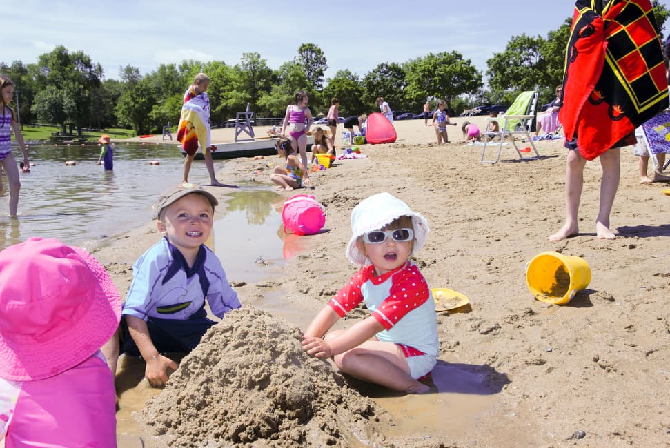 A close up of two toddlers playing in the sand at Lake Taghkanic State Park, with several other families along the beach in the background.