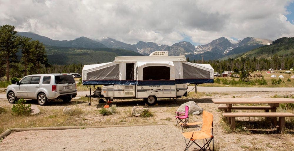 Pop-up camper being towed by a large vehicle sits at a campsite within the mountains. 