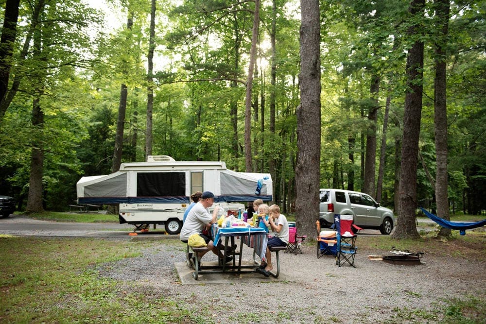 Large family sits at a picnic table enjoying a meal with their pop-up camper in the background.