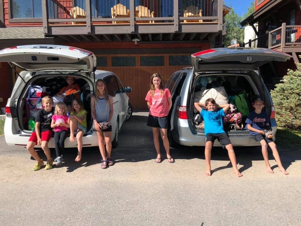 7 kids stand in front of 2 packed vans in preparation of a road trip. Setting up your car is the next step to staying sane on family road trips.