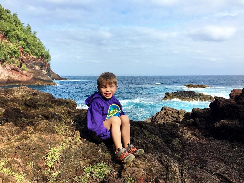 Little boy sits on a rock in front of the beach in Maui, one of the best hot places to visit in December with kids!