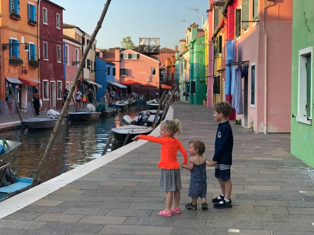 Three young children stand looking at a canal in the colorful island of Burano, Italy.