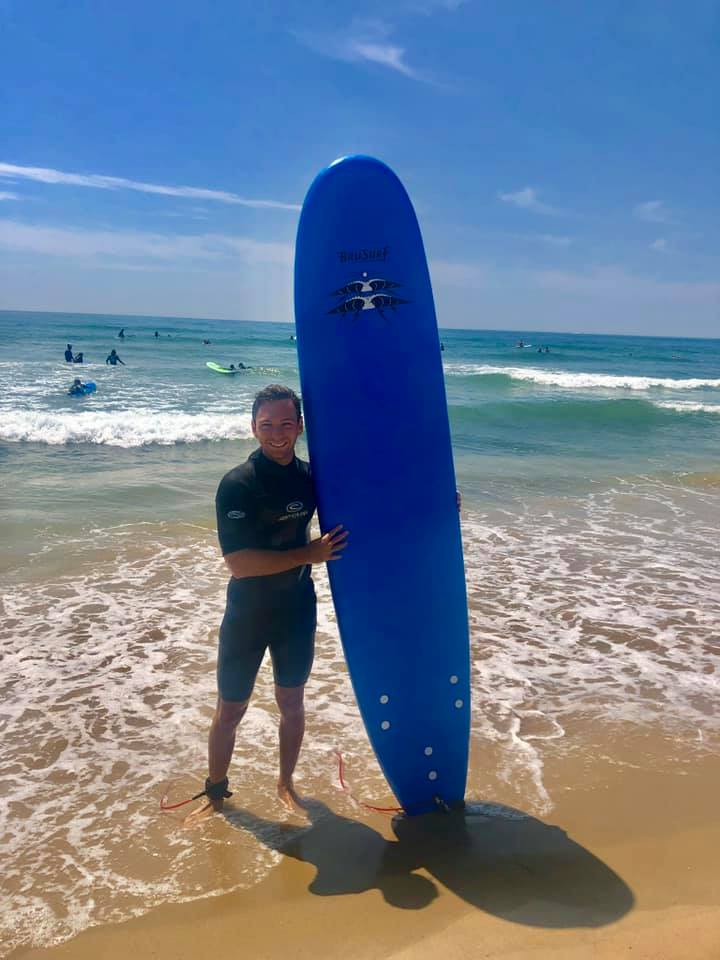 A man holds a large blue surfboard while standing in the water at Montauk Beach.