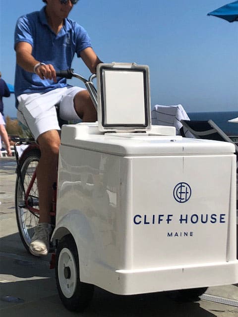 A staff member bikes around the property at Cliff House Maine, with pushing an ice cream cart.