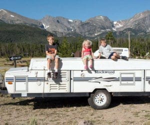 Three kids sitting on an RV pop-up trailer in front of mountains