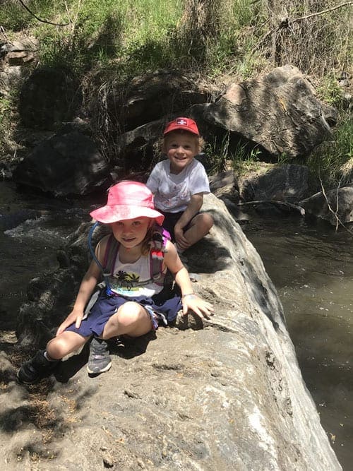 Two young children sit on a log over a stream while hiking near Denver.