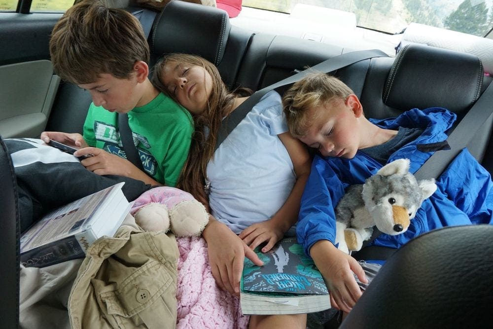 Three kids sit in the back of the car, two sleep while the third reads, great ways for Staying Sane Family Road Trips.