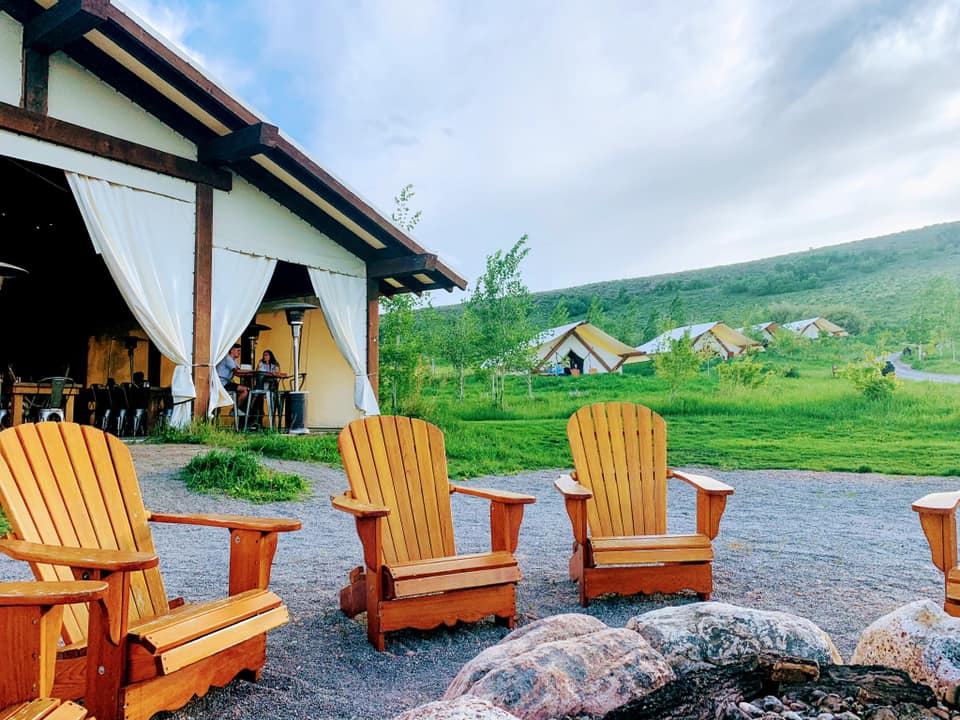 Three adirondack chairs sit by a fire pit in front of a luxury tent.