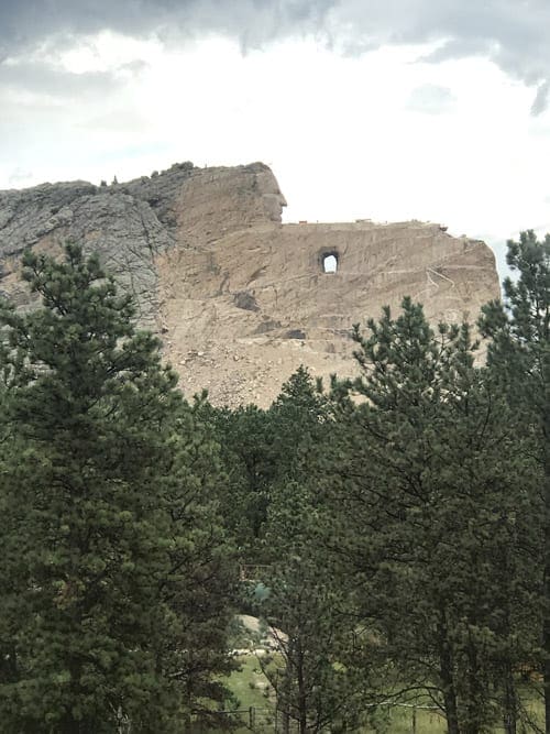 A view of the Crazy Horse Memorial in the distance with evergreen trees lining the foreground.