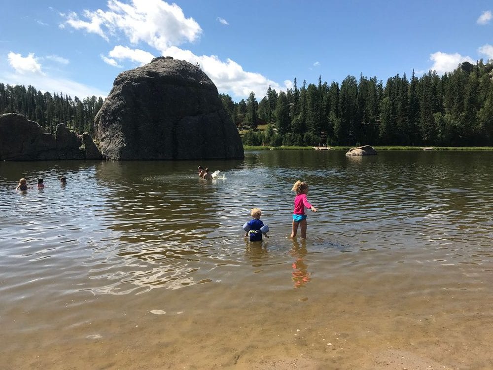 Two young kids play in a lake with a large bolder and row of evergreen trees in the background on the grounds of Custer State Park.