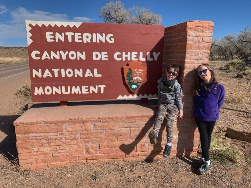 Two kids stand happily in front of the entrance sign for Canyon De Chelly National Monument.