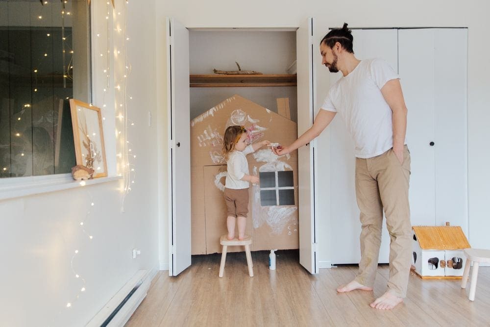 Father and daughter decorate a cardboard fort with white paint. Creating an indoor jungle to explore is a unique family staycation idea.