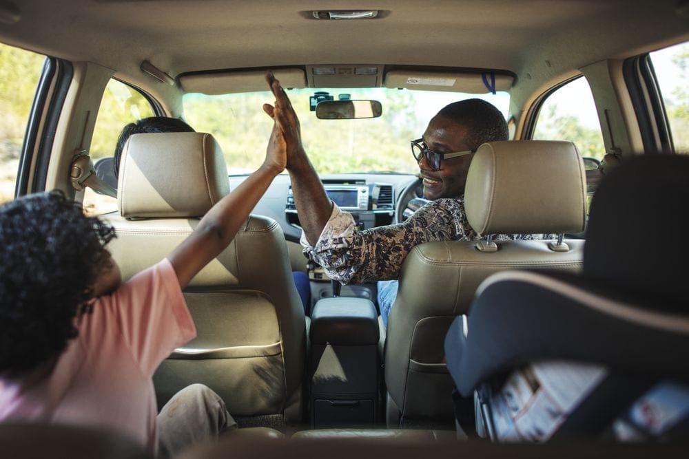 Inside a car, father and child highfive while the other parent drives.