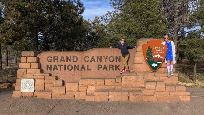 Two kids stand happily in front of the entrance sign for Grand Canyon National Park.