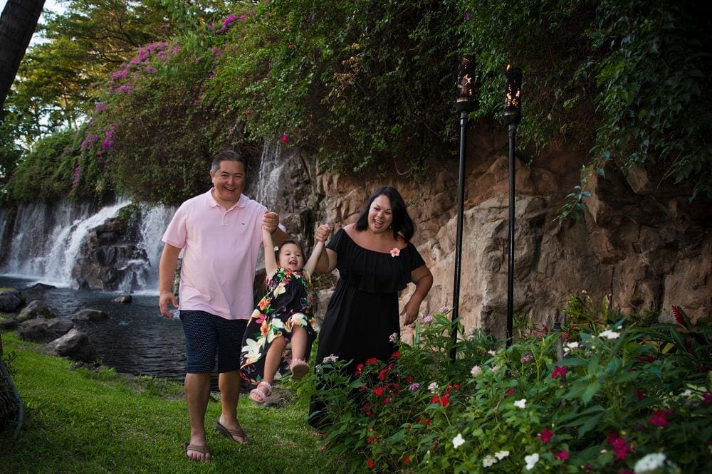 Parents swing their young daughter while walking in a lush Hawaiian park filled with flowers and a waterfall in the backfound. Flexibility of benefits is another reason to purchase travel insurance for families.