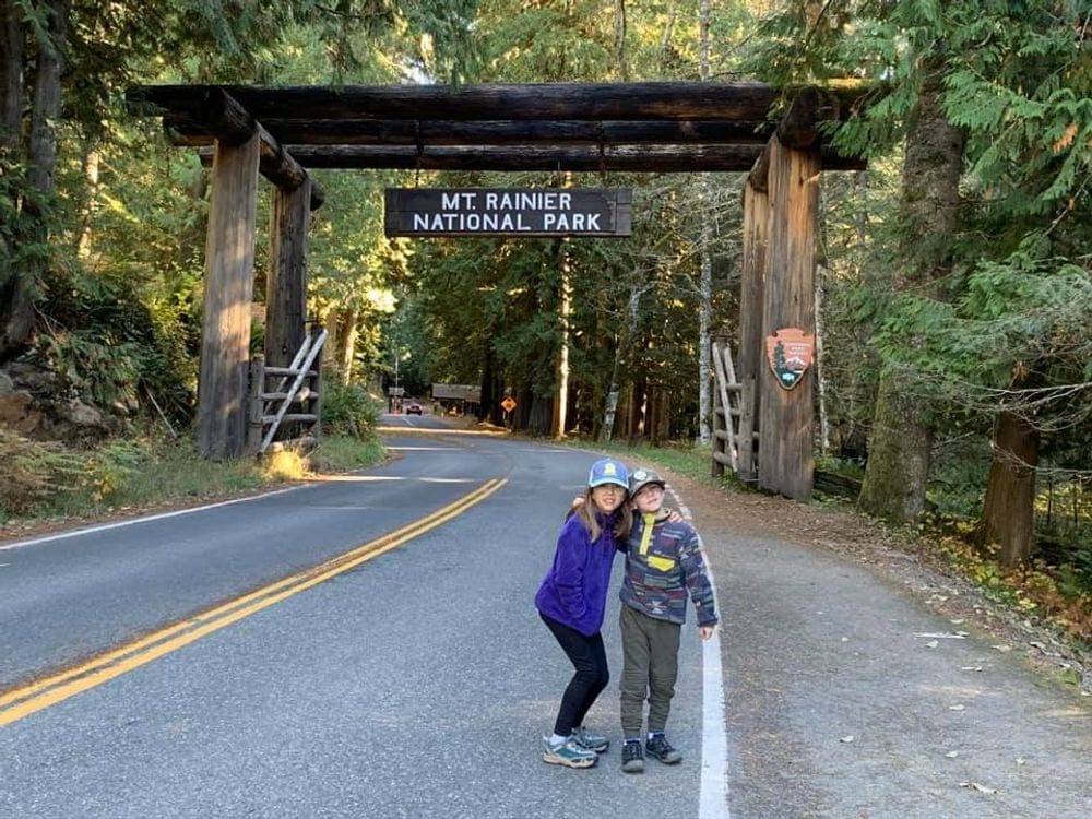 Two young kids stand in front of the entrance sign at Mt. Rainier National Park.