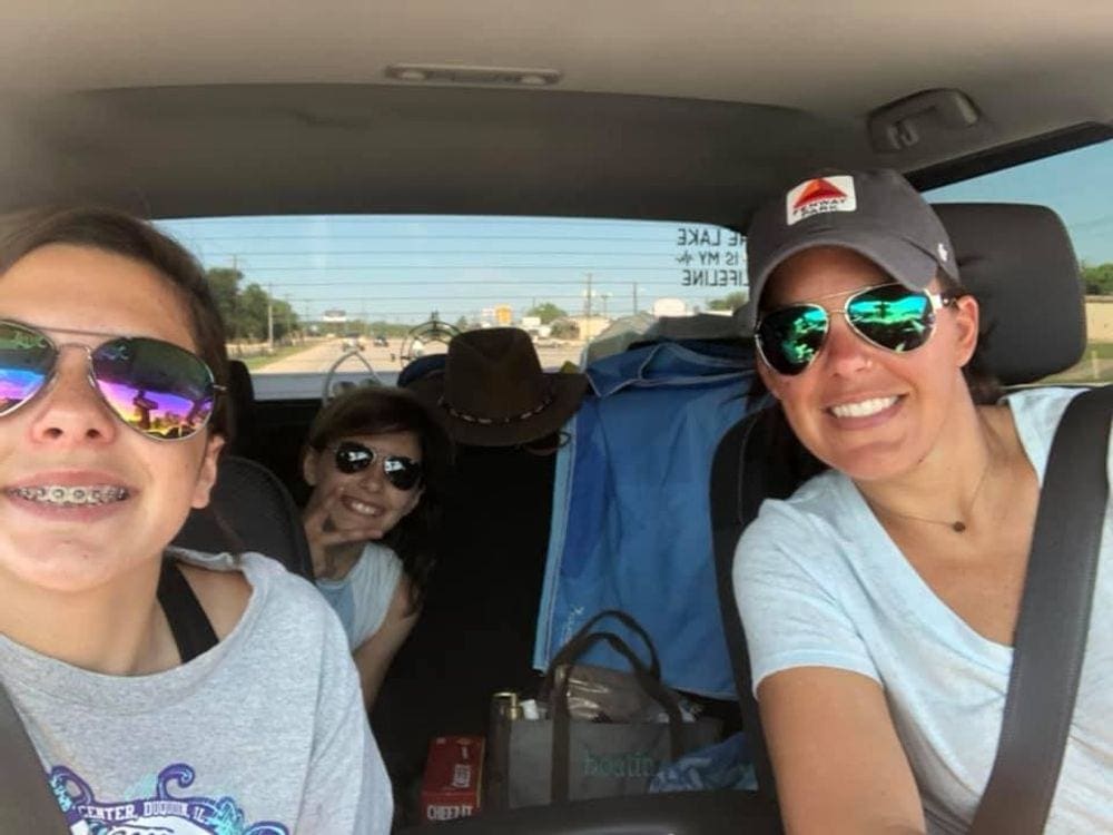 A mom and her two daughters smiling in the car, all wearing matching sunglasses.