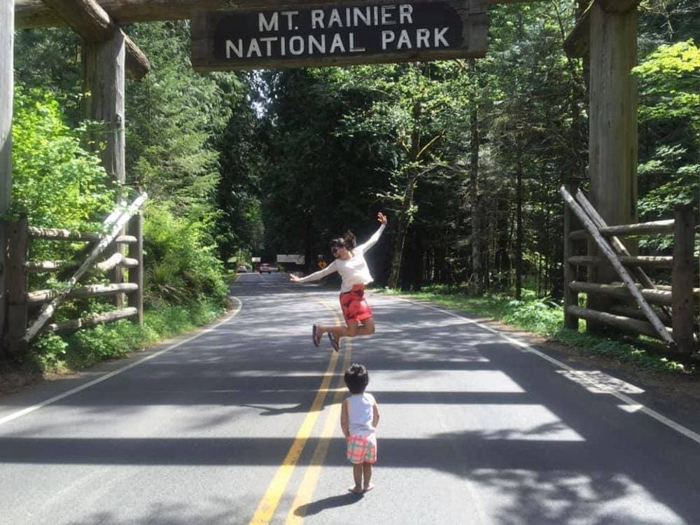 A mom jumps in front of the Mt. Rainier National Park sign while her toddler son looks on.