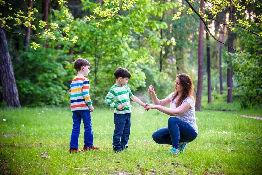 Mom spraying two boys with bug repellent in the woods. Preparing for local insects is one of our tips for traveling with children who have allergies.