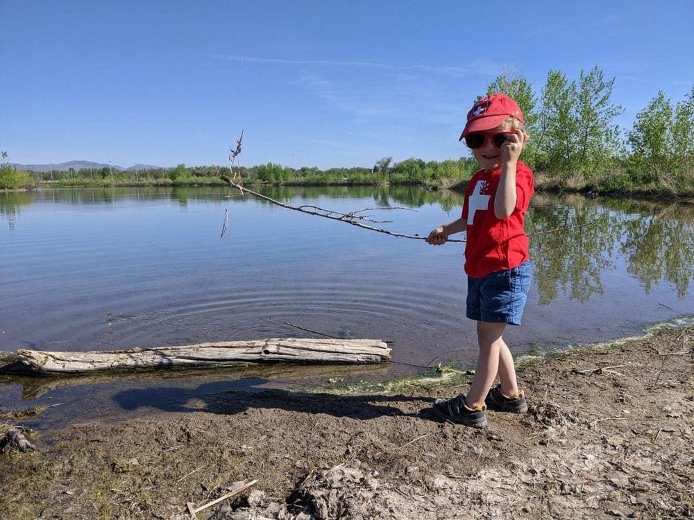 A young boy holds his sunglasses to his face in one hand and a large stick in the other in front of a large river with trees standing along the opposite bank.